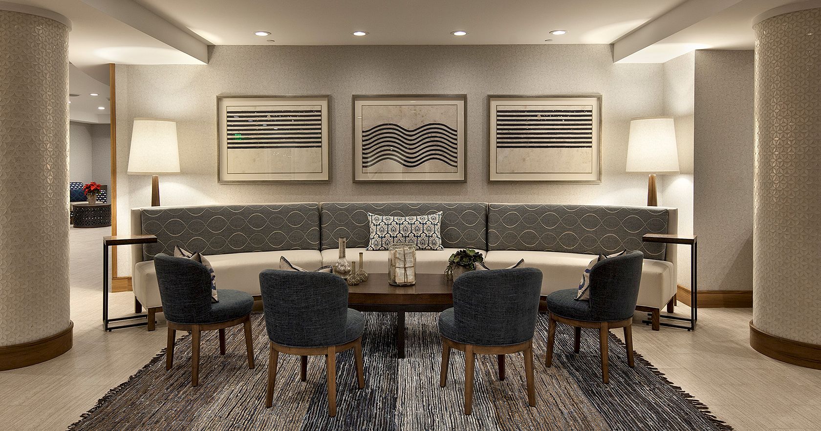 A modern hotel lounge with a curved sofa, six dark chairs around a round table, muted lighting, two lamps, abstract wall art, and a patterned rug.