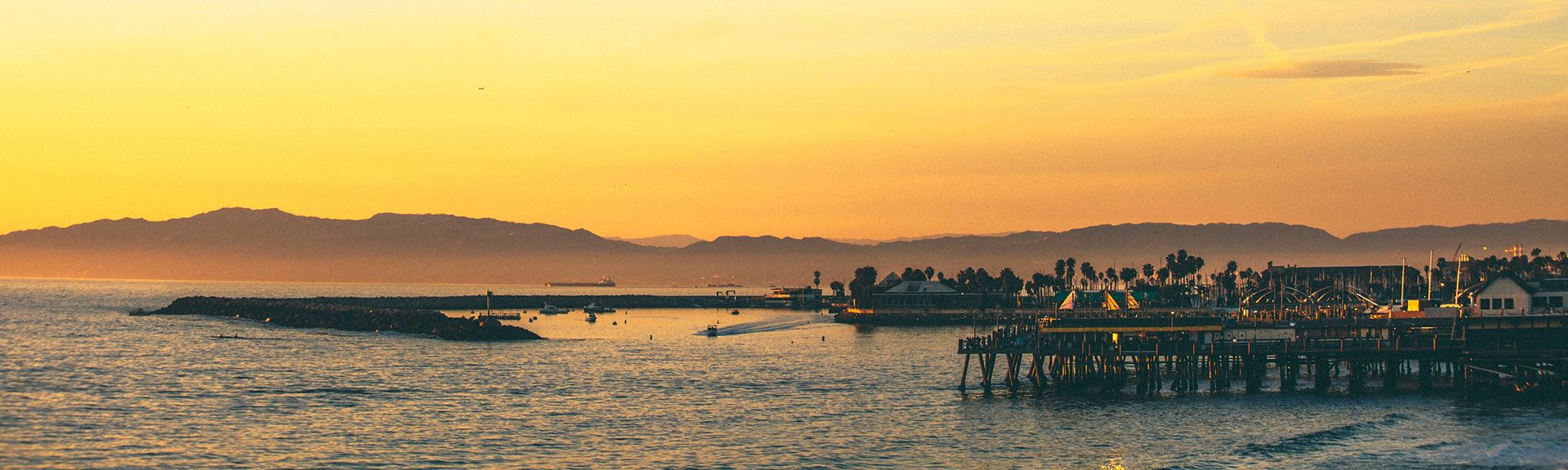 Sunset over a calm harbor with a pier, distant hills, and silhouettes of boats and palm trees along the shoreline.