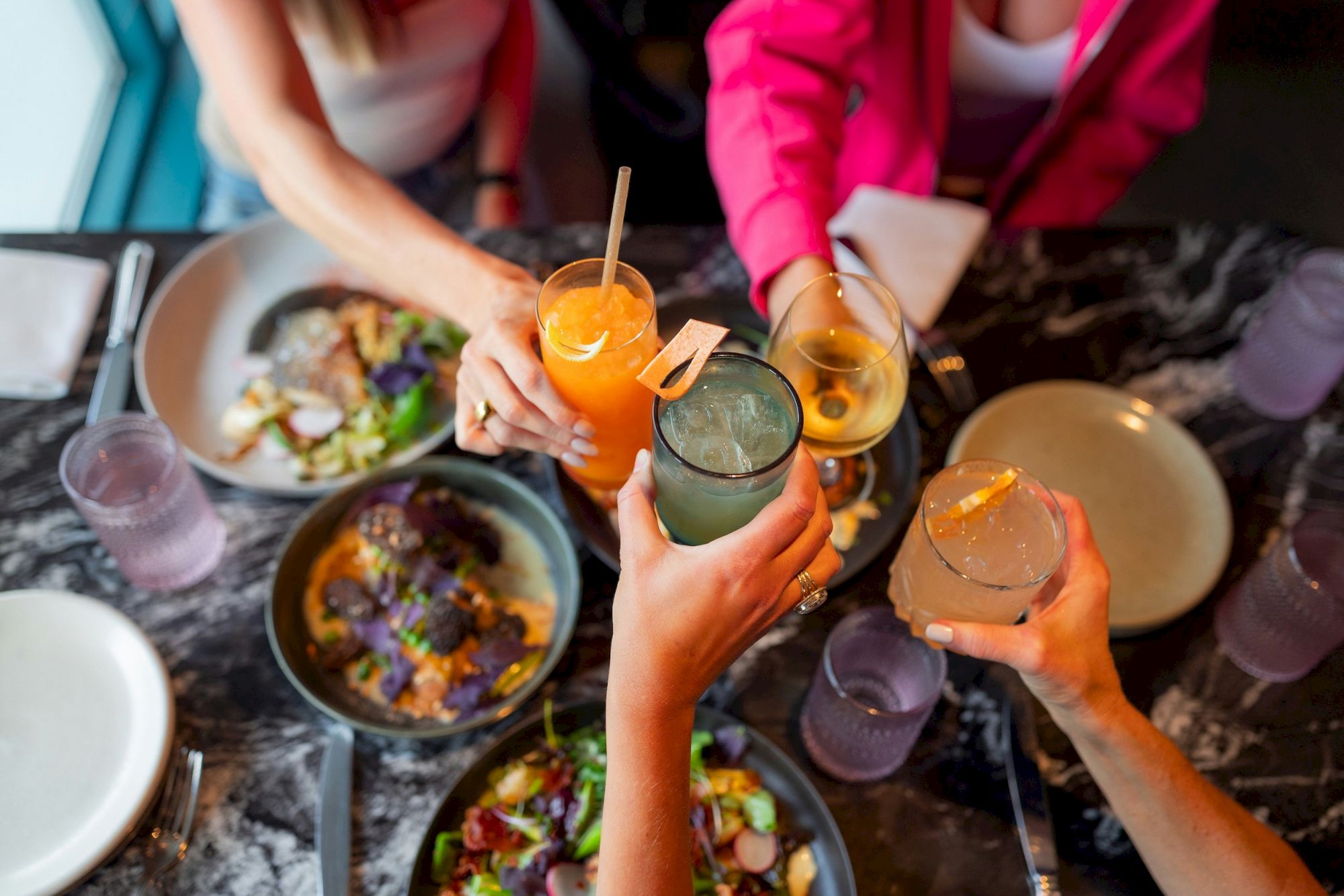 A group of friends cheers with colorful cocktails over a spread of bowls and plates at a social meal.
