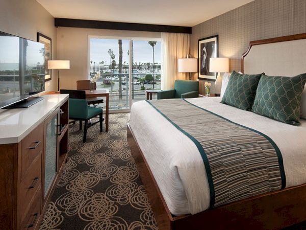 A modern hotel room with a king bed, desk, flat-screen TV, and a balcony overlooking a marina, bright lighting and patterned carpet.