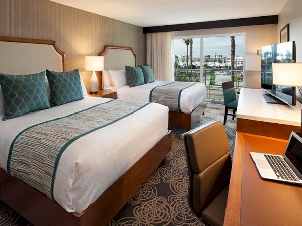 Two clean hotel beds, a desk with laptop, a TV, and a window cityscape view; cozy lamps and teal accents tie the room together.