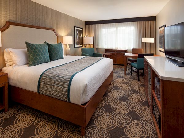 A hotel room with a large bed, teal accents, bedside lamps, a work desk, a TV, and a patterned carpet, bright window light, tidy and welcoming.