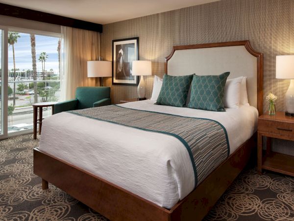 A luxurious hotel room with a large bed, teal accents, two nightstands, lamps, a seating chair, and a view of palm trees through a window, ending this sentence.