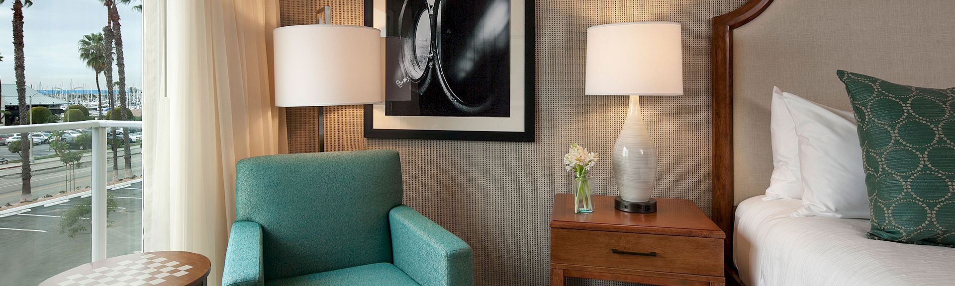 A stylish hotel room with a teal armchair, wooden nightstand, white lamps, framed art, and a bed with green patterned pillow, near a window.