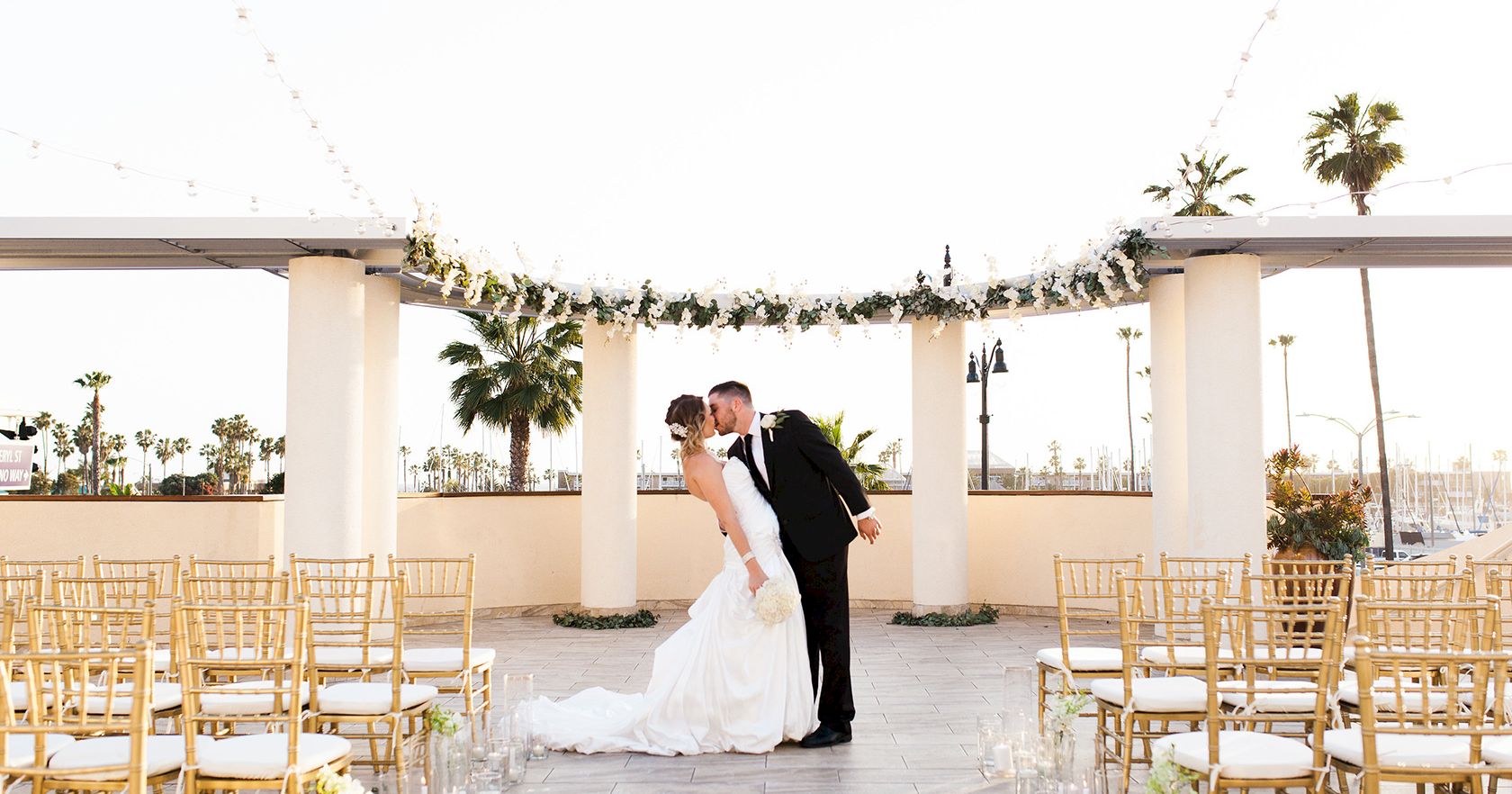A bride and groom kiss at an outdoor wedding altar, surrounded by gold chairs and hanging garlands, on a sunny terrace.