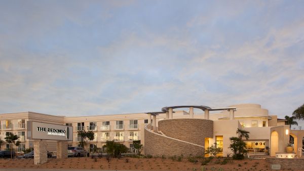 A wide, low-rise hotel with a curved entrance, neutral beige tones, palm trees, and a sign reading &ldquo;The Red... [hotel name]&rdquo; under a blue, partly cloudy sky. (End with period.)