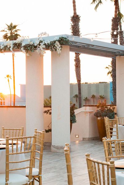 Elegant rooftop wedding setup with white drapes, gold chairs, round tables, floral garlands, and sunset backdrop. A chic outdoor ceremony vibe.