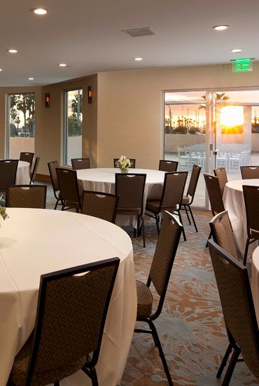 A banquet hall with round tables covered in white linens, each adorned with small floral centerpieces, and soft warm lighting by windows and doors.