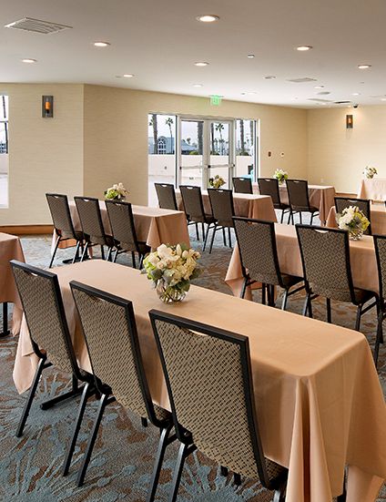 A conference or banquet room set up with long tables draped in peach-tan cloth, floral centerpieces, and chairs arranged for a meeting or event. top it at 140 characters, always ending the sentence.