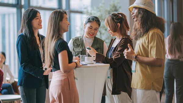 A group of five young adults chat and laugh around a tall white table in a bright, modern space, sharing drinks and smiles.