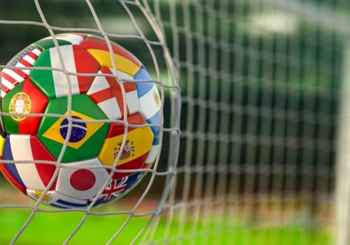A soccer ball with multiple country flags on it gets blocked by a goal net, ready to score on a green field.