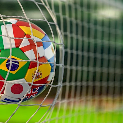 A soccer ball with multiple country flags on it gets blocked by a goal net, ready to score on a green field.