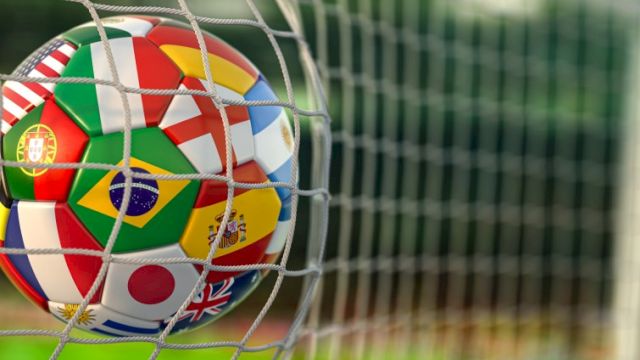 A soccer ball with multiple country flags on it gets blocked by a goal net, ready to score on a green field.