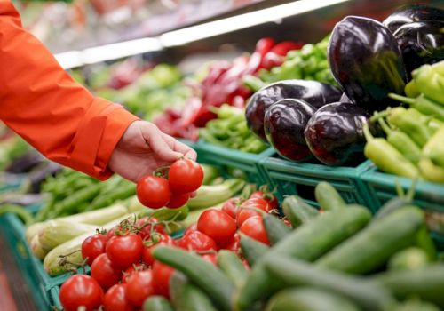 A shopper selects small red tomatoes from a vibrant produce stand with peppers and eggplants nearby, bright market colors all around.