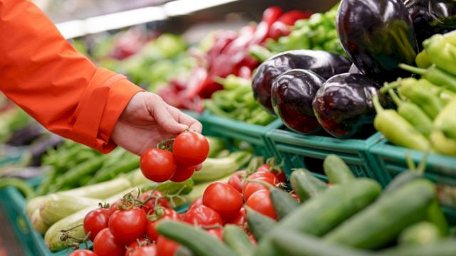A shopper selects small red tomatoes from a vibrant produce stand with peppers and eggplants nearby, bright market colors all around.