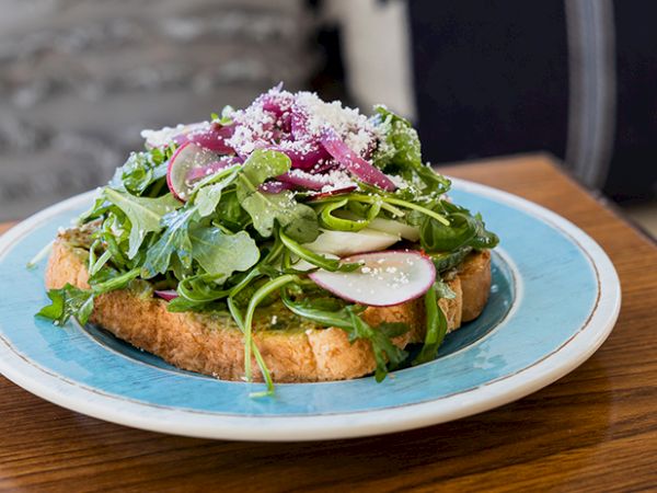 A crispy toast topped with arugula, radish slices, microgreens, shredded cheese, and a tangy dressing on a blue-edged plate.