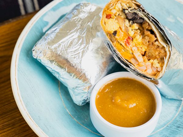 A foil-wrapped burrito halves open to reveal a colorful filling, served with a small cup of brown sauce on a light blue plate.