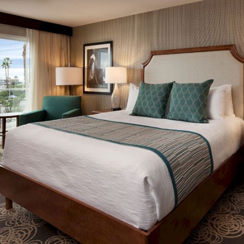 A modern hotel room with a large bed, teal accents, two nightstands, lamps, a chair by the window, and a view outside.