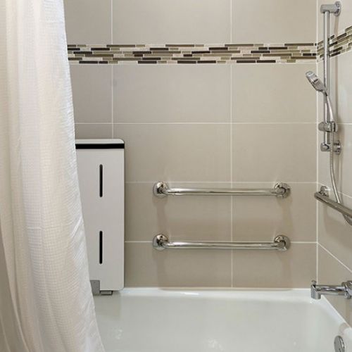 A white bathtub with a curved shower curtain, a vertical white cabinet, and chrome grab bars on the tiled wall, plus a handheld showerhead setup, all in a modern bathroom vibe.