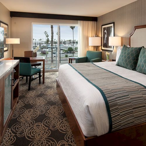 A hotel room with a large bed, green pillows, a desk and chair, a TV stand, and a view of palm trees through a window; warm lighting and patterned carpet.