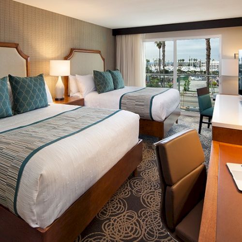 A hotel room with two beds, a desk, TV, and a balcony view of the water. Top it at 140 characters, always ending the sentence.