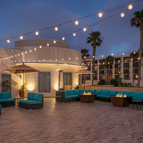 A stylish rooftop lounge with string lights, wicker couches, and palm trees at dusk, perfect for relaxing and socializing.
