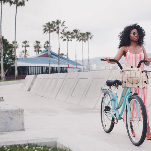 A stylish woman in a pink dress with a pastel bike along a sunny seaside boardwalk. She rides a turquoise bike with pink accents. top it.