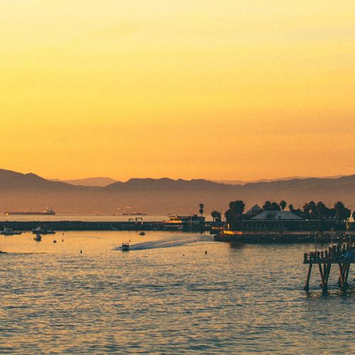 Sunset over calm sea with boats near the shore, warm orange and gold sky reflecting on gently rippling water, peaceful coastal scene.