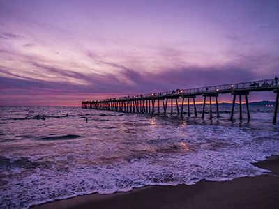 A long wooden pier stretches into the ocean at sunset, purple-pink sky, gentle waves washing onto the sandy beach, calm and serene.