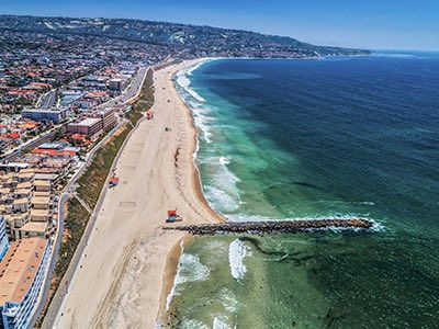 Aerial view of a long sandy beach with turquoise water, a pier jutting into the waves, and a coastal town of low-rise buildings along the shore.