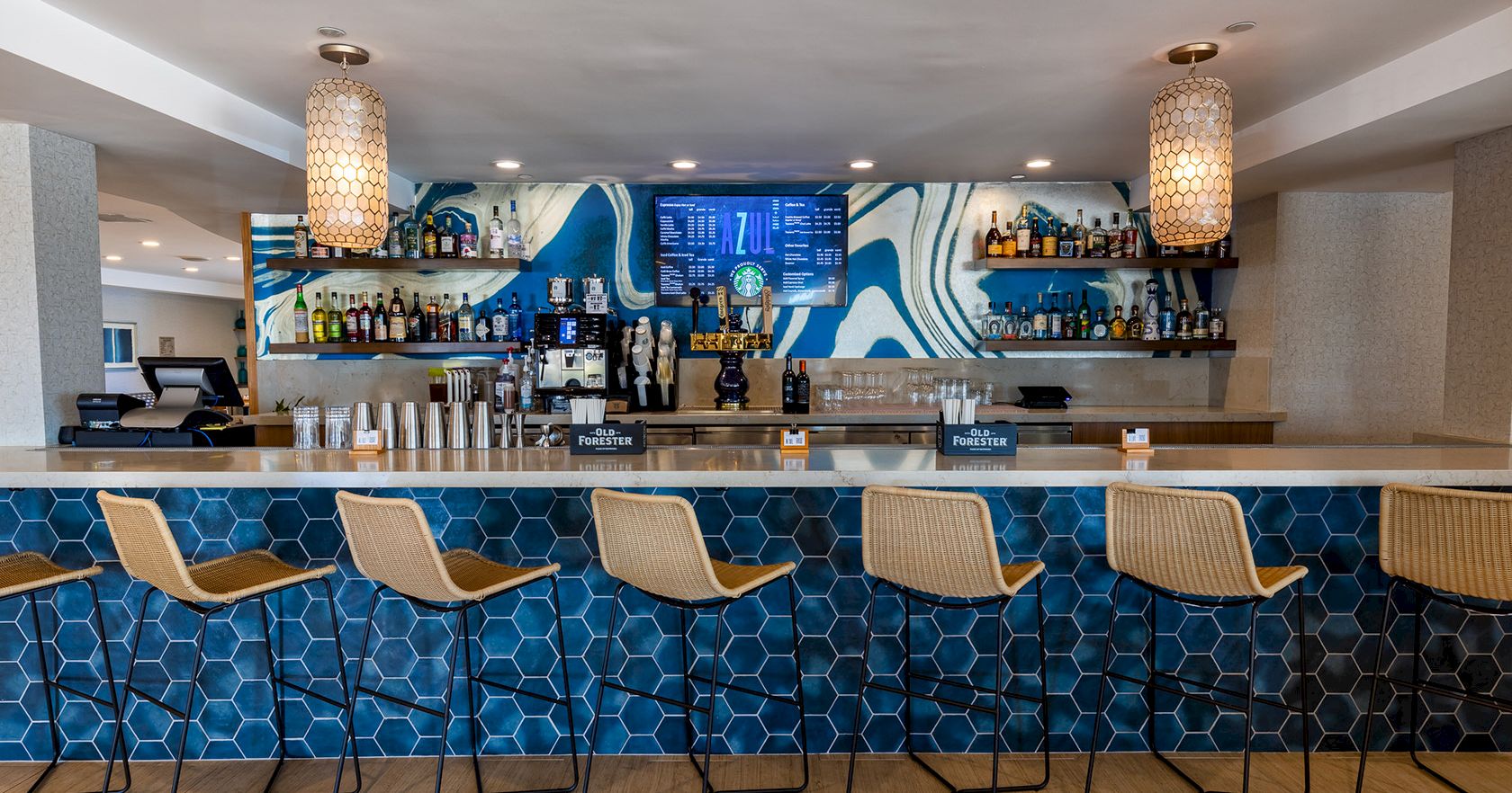 A stylish beach bar with a blue tiled front, eight wicker stools, a long counter, and bottles on glass shelves behind, under pendant lights. The scene is bright and modern.