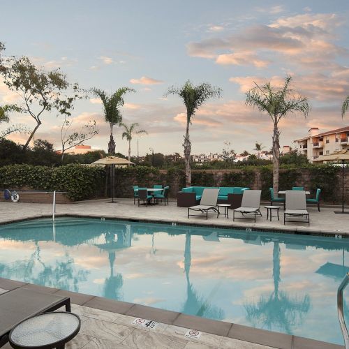 A calm hotel pool area with lounge chairs, palm trees, and a clear sky reflecting on the water, surrounded by a fence and buildings.