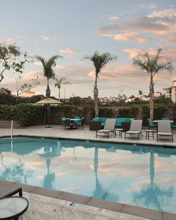A calm hotel pool area with lounge chairs, palm trees, and a clear sky reflecting on the water, surrounded by a fence and buildings.