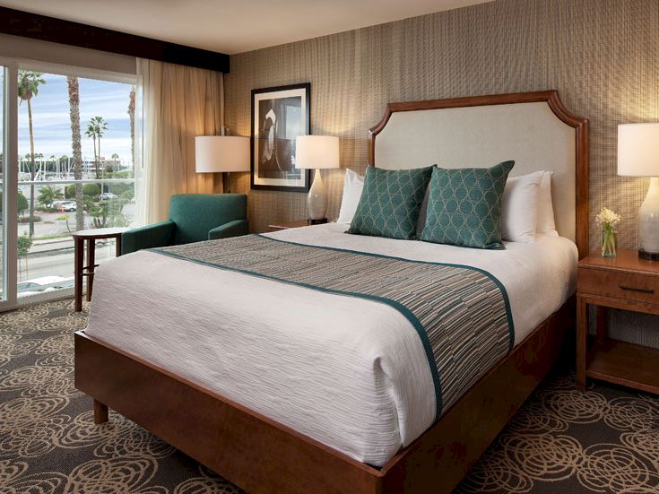 A stylish hotel room with a large bed, teal accents, two bedside lamps, a framed photo, a seating area by glass doors, and ocean-facing views outside.
