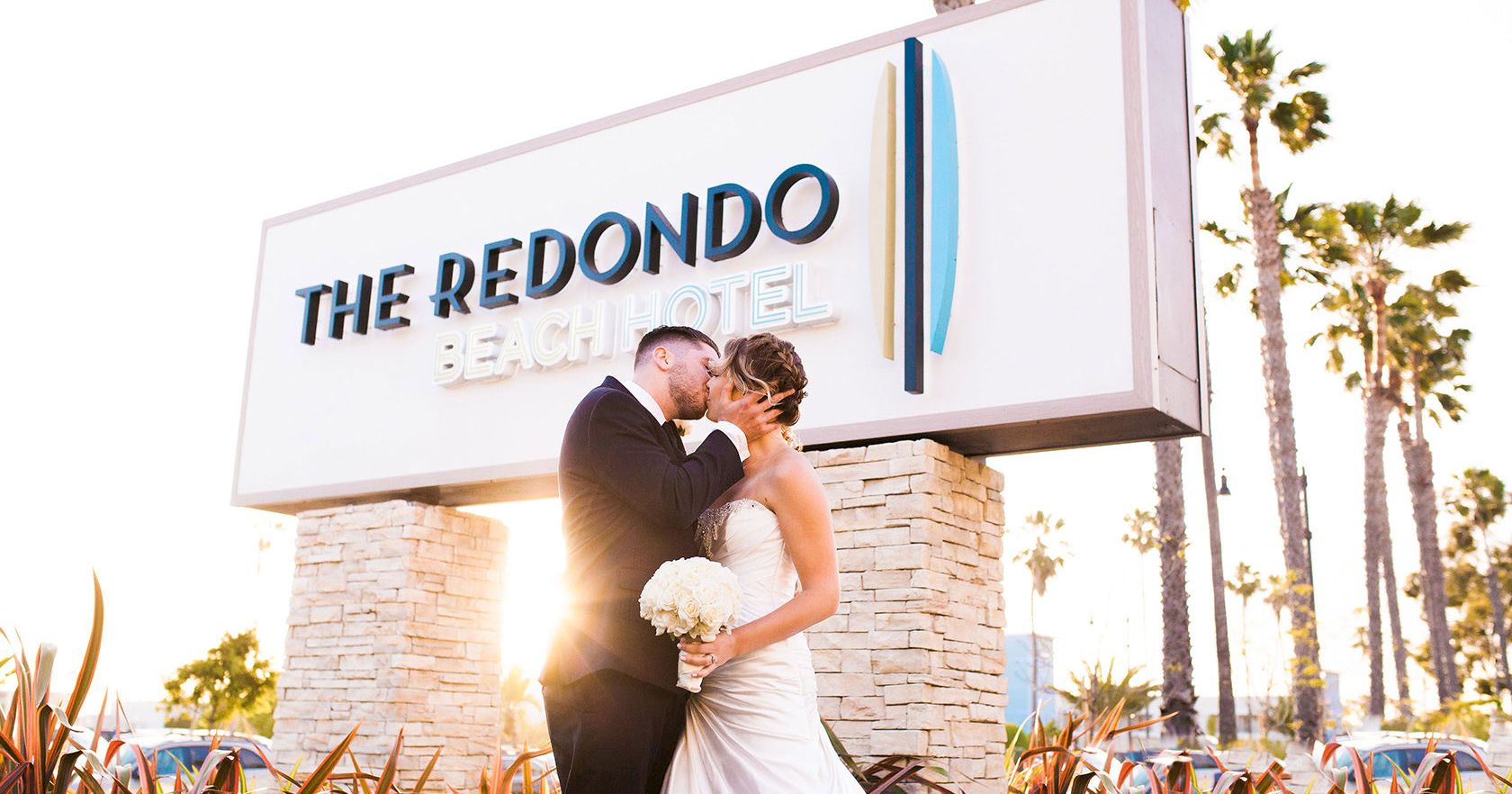 Two newlyweds kiss in front of The Redondo Beach Hotel sign, bride in a white gown holding a bouquet, sunny palm-lined backdrop.