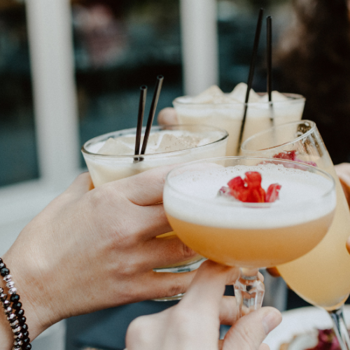 A group of friends toasting with cocktails on a sunny day, glasses raised and smiles around the table.