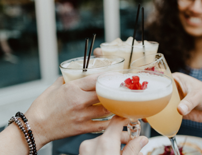 A group of friends toasting with cocktails on a sunny day, glasses raised and smiles around the table.