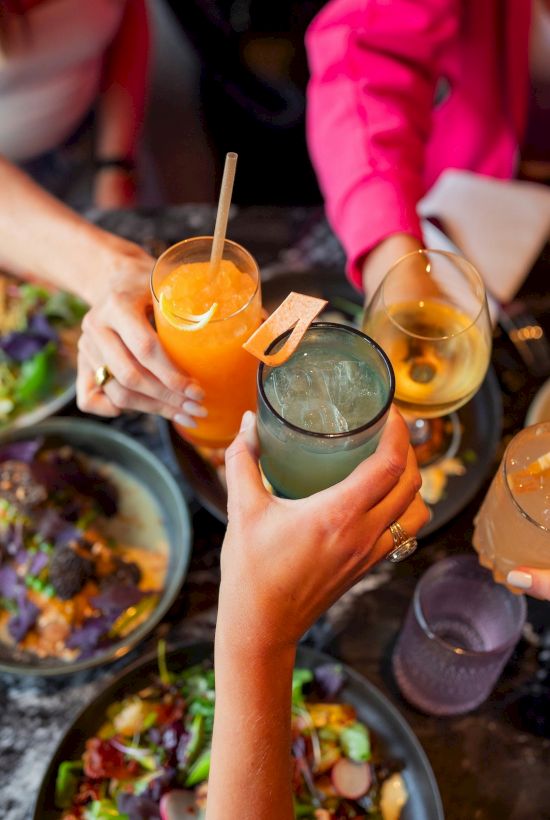 A group cheers with colorful cocktails over a shared table of bowls and plates, celebrating together.